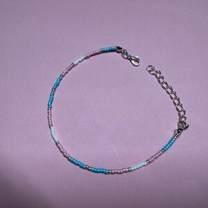 May include: A beaded bracelet featuring a combination of blue, pink, and white beads. It has a silver-coloured chain and clasp. The small beads are evenly spaced, forming a delicate and colourful accessory. The bracelet is approximately 18 cm in length.