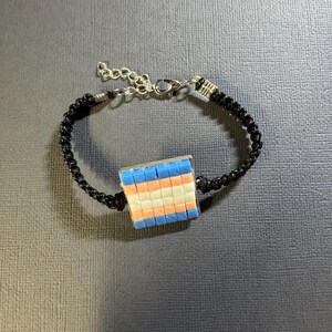 May include: A bracelet featuring a black cord band and a central square charm composed of small, grid-patterned tiles in horizontal stripes of light blue, white, and light pink. The bracelet includes a silver-toned metal lobster claw clasp.