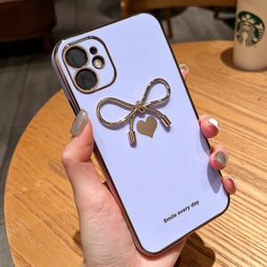 Puede incluir: Funda de teléfono lila con un lazo y un corazón dorados. La funda tiene un borde y un contorno de cámara dorados. El texto "Smile every day" está impreso en la parte inferior. Diseñado para un smartphone.