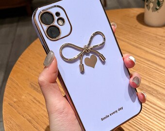 Op de afbeelding: Een lavendelkleurige telefoonhoes met een goudkleurige strik en hartontwerp. De hoes heeft een goudkleurige rand en cameraring. De tekst "Smile every day" staat onderaan. Ontworpen voor een smartphone.