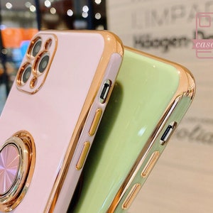 May include: Close-up of two phone cases. One is light pink with a gold ring holder and gold trim. The other is light green with gold trim. The cases have a glossy finish and are designed to protect the phone.