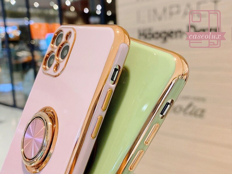 May include: Close-up of two phone cases. One is light pink with a gold ring holder and gold trim. The other is light green with gold trim. The cases have a glossy finish and are designed to protect the phone.
