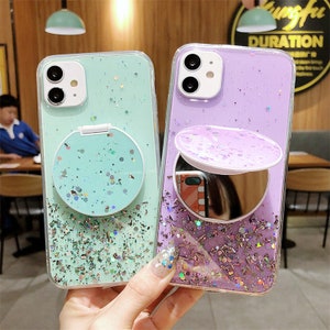 Puede incluir: Dos fundas transparentes para teléfono con purpurina y espejo. Una funda es verde menta, la otra es lavanda. Ambas tienen un agarre emergente redondo lleno de purpurina. La funda lavanda tiene un pequeño espejo.