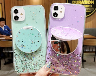 Op de afbeelding: Twee transparante telefoonhoesjes met glitter en een spiegel. Eén hoesje is mintgroen, de andere is lavendel. Beide hebben een ronde, met glitter gevulde pop-up grip. Het lavendel hoesje heeft een kleine spiegel.