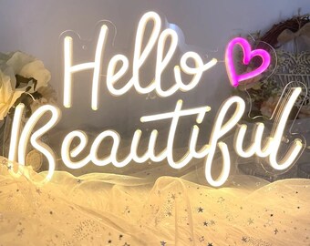 Op de afbeelding: Een neonbord met de woorden "Hello Beautiful" in een sierlijk handschrift, verlicht met een warm wit licht. Een roze hartvorm bevindt zich boven het woord "Beautiful". Het bord is bevestigd op een transparante achtergrond, tegen een zachte stof.