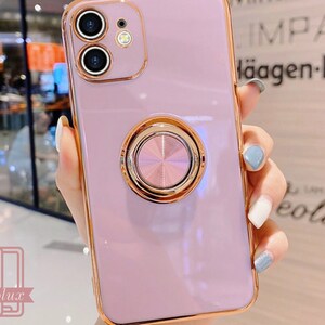May include: A lavender phone case with a gold-colored frame and a matching ring holder. The case has a dual-camera design and the brand name "caselux" is visible in the lower left corner.