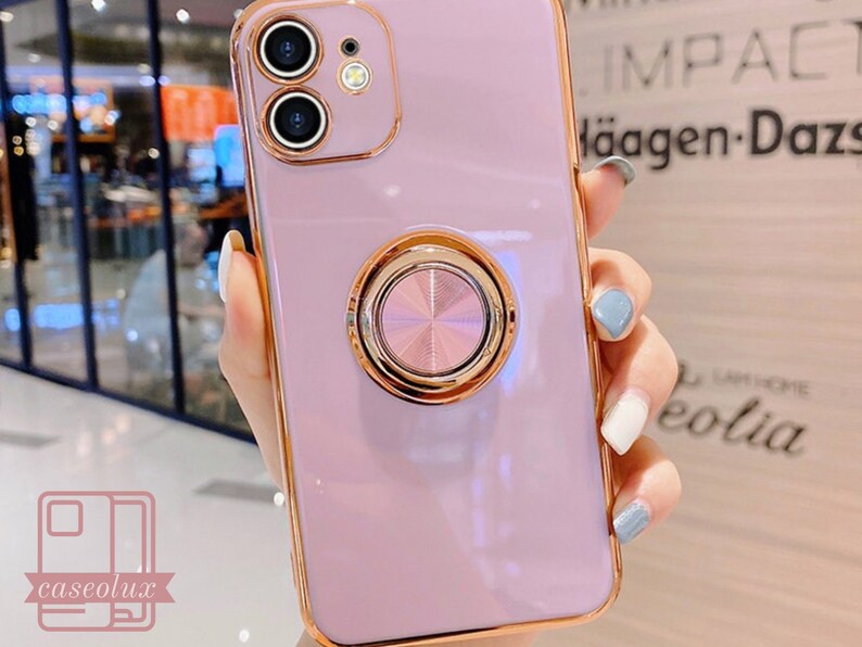 May include: A lavender phone case with a gold-colored frame and a matching ring holder. The case has a dual-camera design and the brand name "caselux" is visible in the lower left corner.