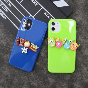 Peut inclure: Deux coques de téléphone : une bleue, une verte. La coque bleue présente une bouteille Coca-Cola, un tigre, un chat et un gant blanc, avec le texte "Toy Story". La coque verte a des figurines colorées en forme de monstres.