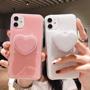 May include: Two glittery phone cases, one pink and one white, each with a heart-shaped pop-up grip. The cases have a clear border and are held up by hands. The pink case has a matching pink heart grip, while the white case has a white grip.