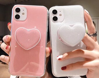 Op de afbeelding: Twee glitterende telefoonhoesjes, één roze en één wit, elk met een hartvormige pop-up grip. De hoesjes hebben een transparante rand. De roze hoes heeft een bijpassende roze hartgrip, de witte een witte grip.