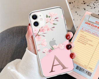 Op de afbeelding: Transparante telefoonhoes met een roze bloemenpatroon en de letter "A". De hoes heeft een roze onderkant en een transparante bovenkant met een bloemenpatroon. De telefoonhoes ligt op een roze klembord.