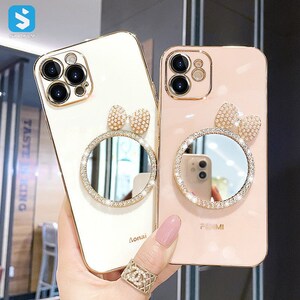 Puede incluir: Dos fundas doradas para teléfono, una crema y otra rosa, cada una con un espejo circular y un adorno en forma de lazo. Los espejos están rodeados de pedrería. La funda crema tiene la palabra "Aonai" impresa.