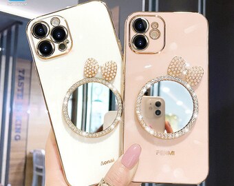 Op de afbeelding: Twee goudkleurige telefoonhoesjes, één crème en één roze, elk met een ronde spiegel en een strikvormige versiering. De spiegels zijn omgeven door strass. Op de crème hoes staat het woord "Aonai".