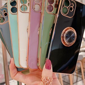 May include: A hand holds a fan of colorful phone cases. The cases are in shades of green, white, purple, and black, with gold-colored trim and camera rings. Each case has a metallic ring holder on the back. The brand name "caseolux" is visible in the lower left corner.