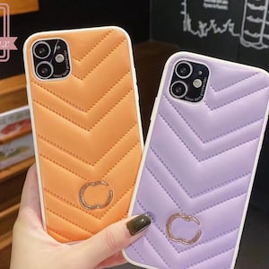 Puede incluir: Se muestran dos fundas para teléfono, una naranja y otra lavanda. Ambas presentan un diseño de chevrón acolchado y un emblema dorado. Las fundas tienen un borde blanco y un protector de lente.