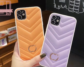 Op de afbeelding: Twee telefoonhoesjes worden getoond, één oranje en één lavendelkleurig. Beide hebben een gewatteerd chevronpatroon en een goudkleurig embleem. De hoesjes hebben een witte rand en een cameralensbeschermer.