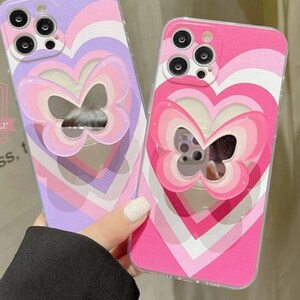 May include: Two phone cases with a butterfly design. The cases feature a heart-shaped cutout with a butterfly-shaped mirror. One case is pink and the other is purple, both with a striped heart pattern.