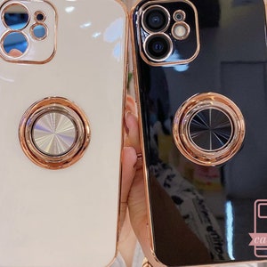 May include: Two phone cases, one white and one black, with a glossy finish and a gold-colored ring holder on the back. The cases have a gold-colored trim around the edges and camera. The white case is on the left, and the black case is on the right.