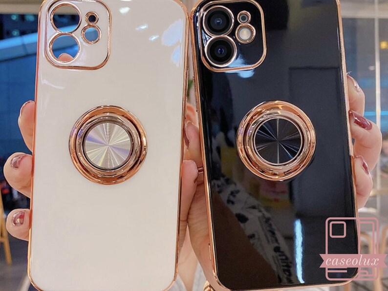 May include: Two phone cases, one white and one black, with a glossy finish and a gold-colored ring holder on the back. The cases have a gold-colored trim around the edges and camera. The white case is on the left, and the black case is on the right.