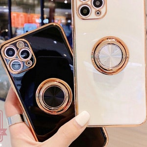 May include: Two phone cases, one black and one white, with gold-colored trim and ring holders. The cases have a glossy finish and are designed to fit a smartphone. The ring holders are circular and provide a secure grip.