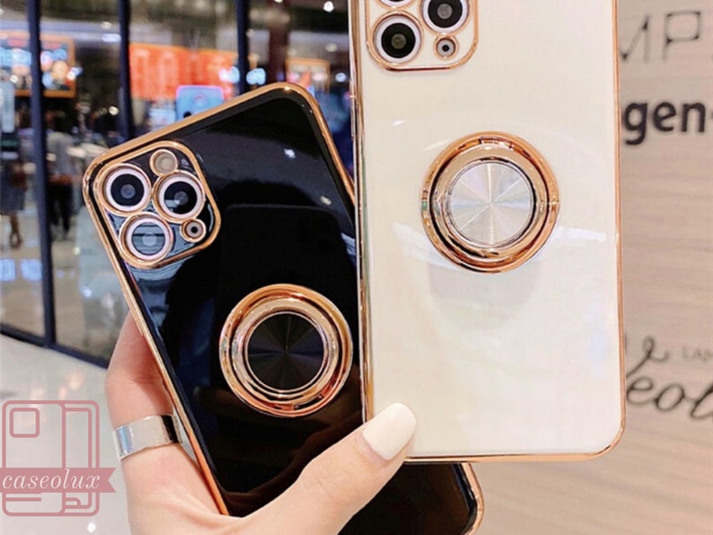 May include: Two phone cases, one black and one white, with gold-colored trim and ring holders. The cases have a glossy finish and are designed to fit a smartphone. The ring holders are circular and provide a secure grip.