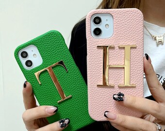 Op de afbeelding: Twee telefoonhoesjes worden getoond, één groen en één roze. Elk hoesje heeft een goudkleurige letter, "T" op het groene hoesje en "H" op het roze hoesje. De hoesjes hebben een getextureerde, leerachtige uitstraling.