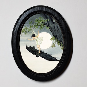 May include: A framed watercolour painting of a fairy riding a bat in front of a full moon. The fairy is wearing a white dress and has brown hair. The bat is black and has large wings. The background is a dark blue sky with a few clouds.