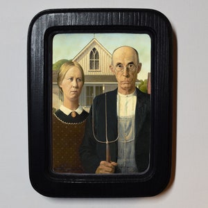 May include: A framed painting of a couple standing in front of a house. The man is wearing a blue jacket and overalls, and the woman is wearing a brown dress with a white collar. The man is holding a pitchfork.