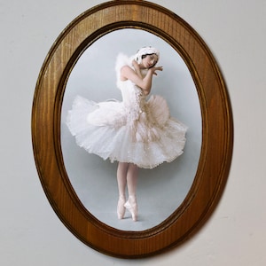 Op de afbeelding: Een ballerina in een witte tutu en pointe schoenen staat in een ovale houten lijst. De ballerina staat geposeerd met haar hoofd naar beneden gekanteld en haar handen voor haar gevouwen.