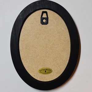 May include: Back of an oval black wooden picture frame with a cardboard backing. A metal hanger is attached to the top center. A small, oval sticker with the text "BLUEBELL ART & WOOD DESIGNS" is affixed to the bottom.