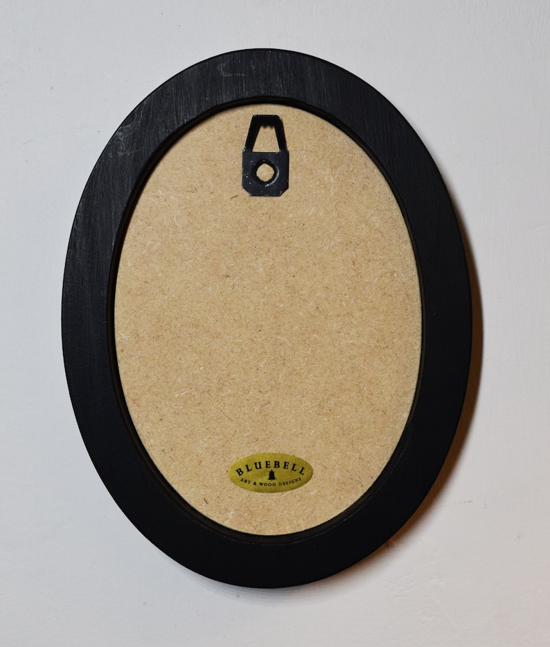 May include: Back of an oval black wooden picture frame with a cardboard backing. A metal hanger is attached to the top center. A small, oval sticker with the text "BLUEBELL ART & WOOD DESIGNS" is affixed to the bottom.