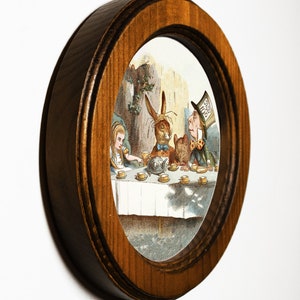 May include: A framed illustration of a tea party scene from Alice's Adventures in Wonderland. The Mad Hatter, the March Hare, and Alice are seated at a table with a teapot and cups. The illustration is in a brown oval frame.
