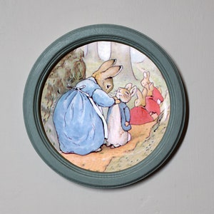 Pode incluir: Uma ilustração emoldurada de um grupo de coelhos em um cenário de floresta. A coelha mãe está vestindo um vestido azul e está ajudando um coelho mais jovem a colocar uma jaqueta. Os outros coelhos estão sentados no chão.