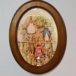 以下が含まれることがあります： ピーターラビットのイラストが描かれた、楕円形の額装されたプリント。プリントには、ピーターラビットと他のウサギが庭園の風景の中に描かれています。フレームは木でできており、茶色の仕上げになっています。