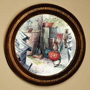 Può includere: Una stampa incorniciata in una cornice rotonda di legno scuro. L'illustrazione raffigura conigli in un giardino, uno dei quali tiene in mano un oggetto rosso. La cornice in legno ha un design decorativo.