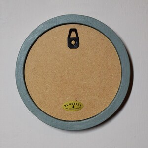 May include: The back of a round, light blue wooden picture frame with a gold sticker that says "Bluebell Art & Wood Designs".