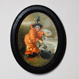 以下が含まれることがあります： 木製のさじで釜を混ぜる魔女のヴィンテージなハロウィーンのイラスト。彼女はとがった帽子と金色の星がついた赤いローブを着ています。画像は黒い楕円形の額縁に入っています。
