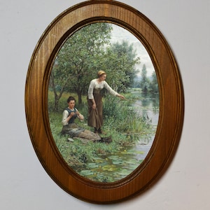 Puede incluir: Una pintura enmarcada ovalada que representa a dos mujeres en un entorno rural. Una mujer está sentada a la orilla de un río, mientras que la otra está de pie y sostiene una caña de pescar. La pintura presenta un exuberante paisaje verde con árboles, agua y nenúfares.