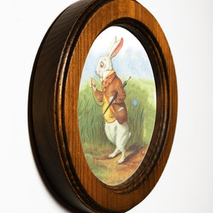May include: A framed illustration of a white rabbit wearing a waistcoat and holding a pocket watch. The rabbit is standing in a grassy field with a blue flower.