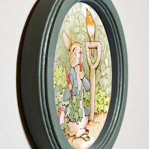 May include: A green oval picture frame with a white oval mat. The picture inside the frame is of a rabbit eating a carrot with a robin perched on a garden tool.