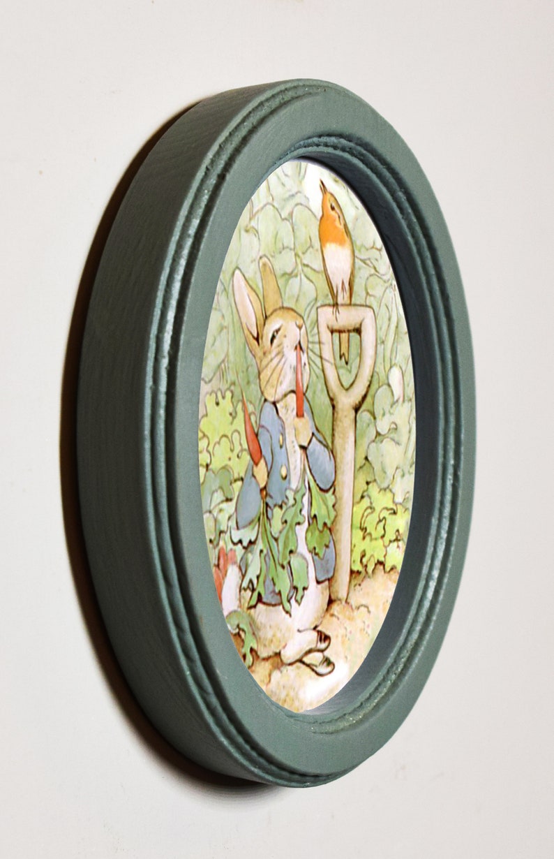 May include: A green oval picture frame with a white oval mat. The picture inside the frame is of a rabbit eating a carrot with a robin perched on a garden tool.