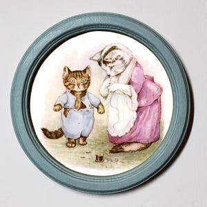 Puede incluir: Una ilustración enmarcada de dos gatos, uno con un vestido azul y el otro con un vestido rosa, con una aguja e hilo en el suelo. La ilustración está en un marco azul claro.