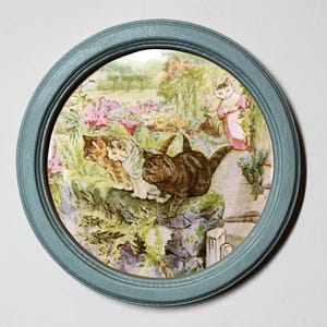 Puede incluir: Un plato redondo de cerámica con un marco azul presenta una ilustración vintage de cuatro gatos en un jardín. Los gatos son marrones, blancos y negros con follaje verde y flores rosas en el fondo.