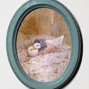 Könnte beinhalten: Eine gerahmte Illustration eines Entchens, das einen blauen Hut und eine rosa-weiße Decke trägt und in einem Nest aus weißen Federn ruht.