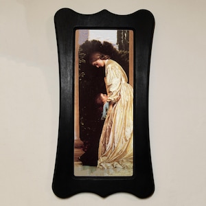 May include: A framed print of a classical painting showing a woman in a gold dress holding a child. The black frame has a decorative, curved design. The artwork depicts a tender moment between the figures.