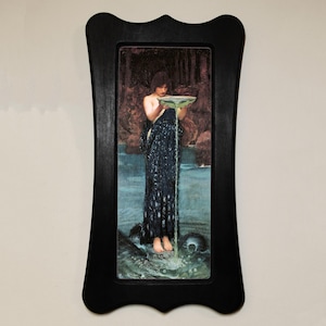 May include: A framed painting depicting a woman in a blue gown pouring water from a bowl. The woman is standing in a body of water with a dark forest in the background.