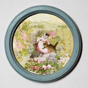 May include: A framed illustration of three cats in a garden setting. The mother cat is wearing a pink dress and holding a kitten. The other kitten is playing with the mother cat's tail. The garden is filled with flowers and greenery.