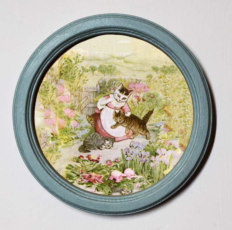 May include: A framed illustration of three cats in a garden setting. The mother cat is wearing a pink dress and holding a kitten. The other kitten is playing with the mother cat's tail. The garden is filled with flowers and greenery.