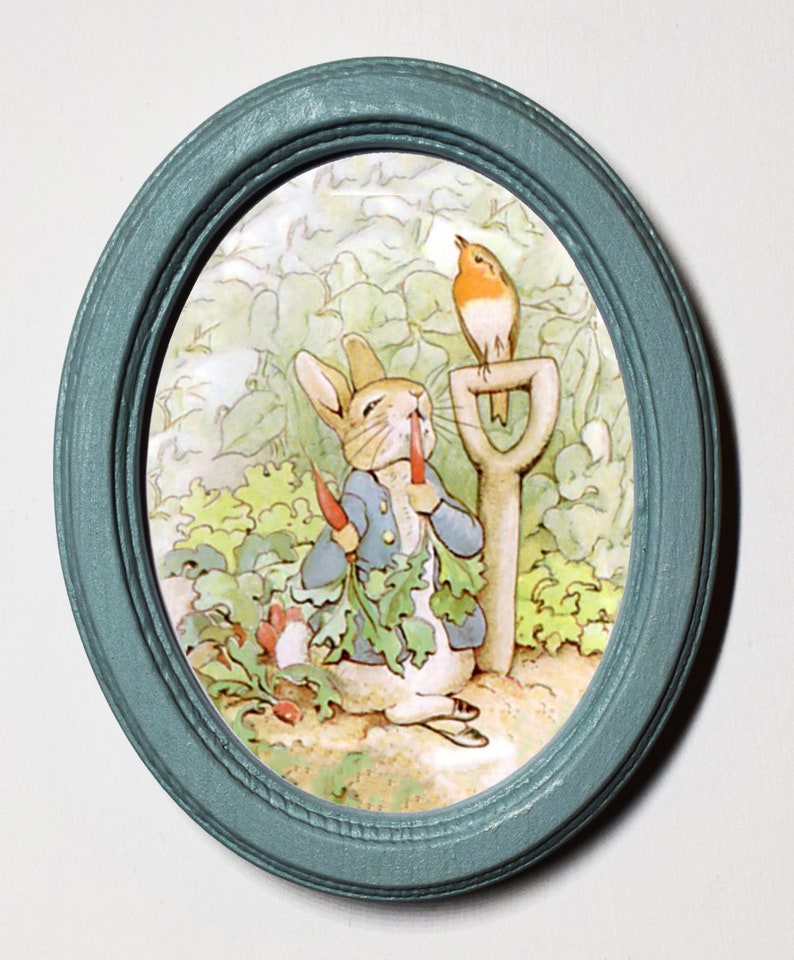 May include: A blue oval frame with a white background featuring a cartoon rabbit holding a carrot and a robin perched on a garden tool. The rabbit is wearing a blue jacket and the robin is red with a brown head.