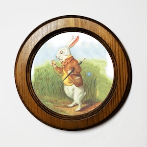 May include: A white rabbit wearing a waistcoat and holding a pocket watch and a cane, is depicted in a vintage illustration on a round wooden coaster.